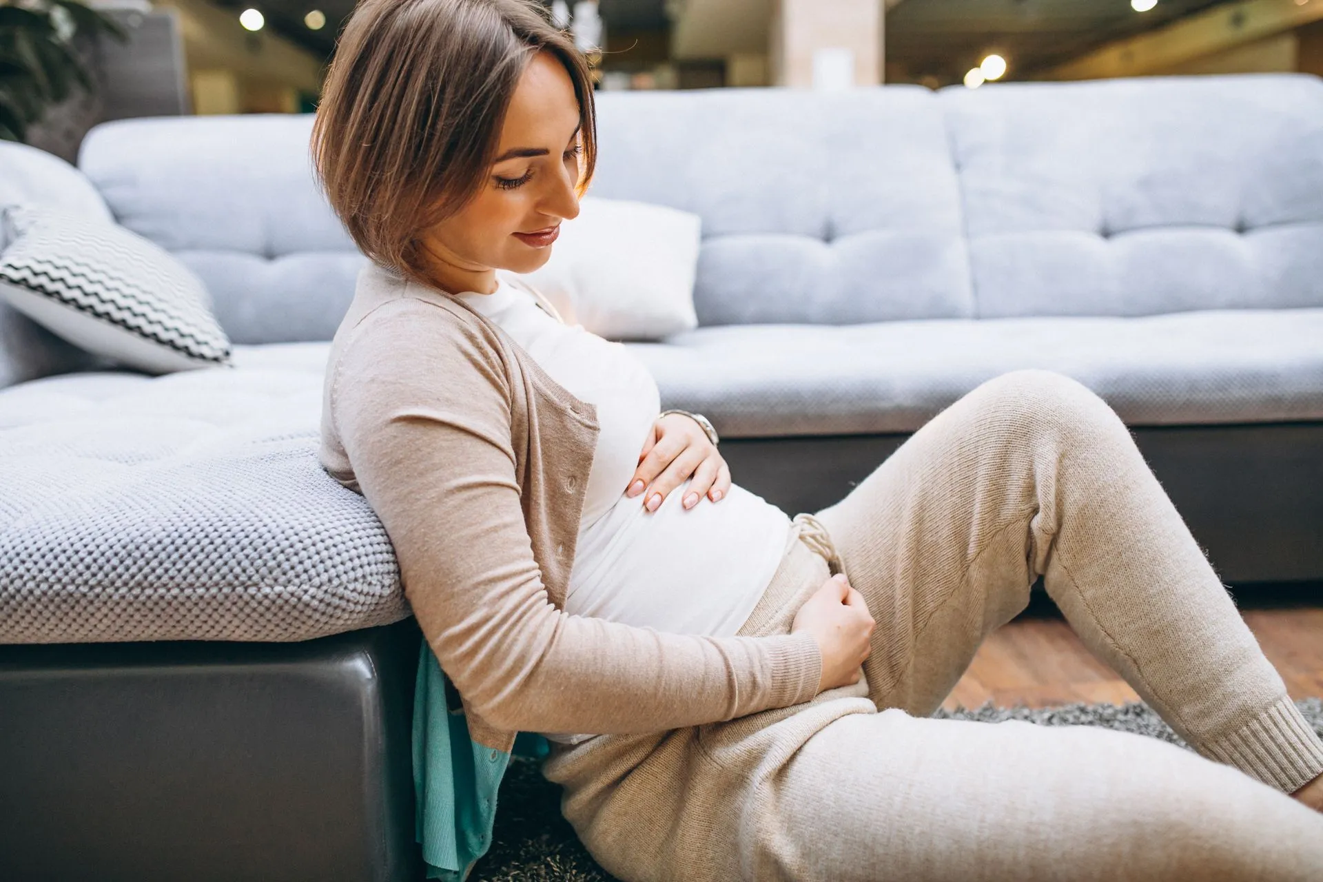 Schwangere Frau sitzt vor dem Sofa und hält ihren Bauch